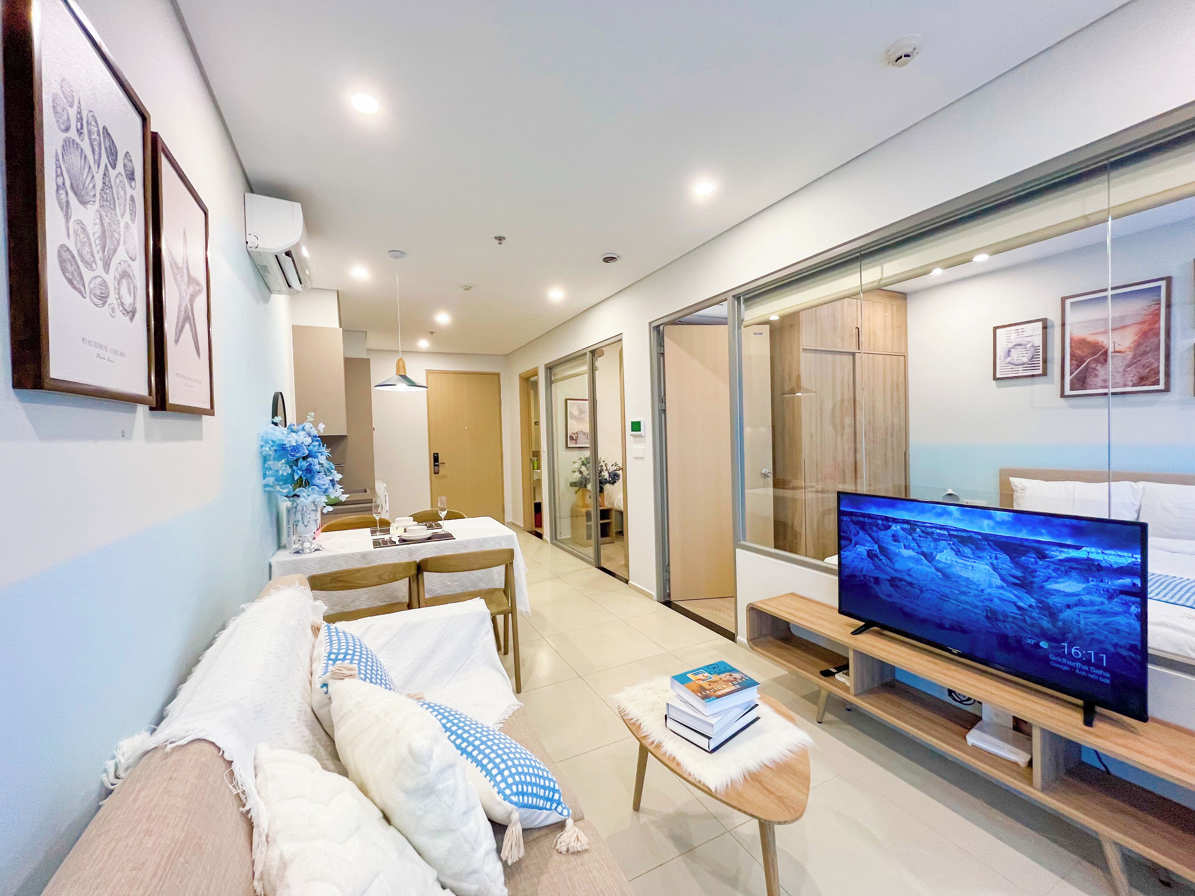 A modern hotel room with a large flat-screen TV, a sofa with cushions, a small table with books, and a sliding glass door leading to a bathroom.