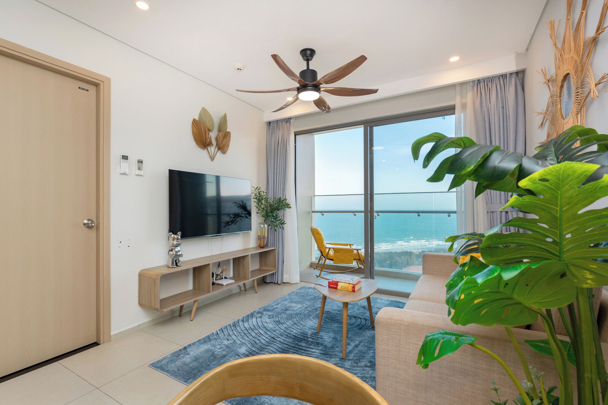 A modern living room with a flat-screen TV, a ceiling fan, a balcony with a view of the ocean, and a large plant.