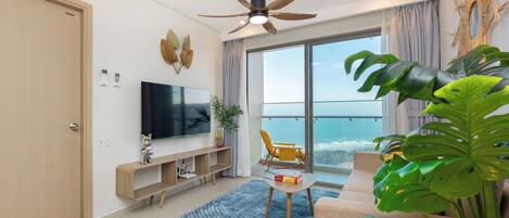 A modern living room with a flat-screen TV, a ceiling fan, a balcony with a view of the ocean, and a large plant.