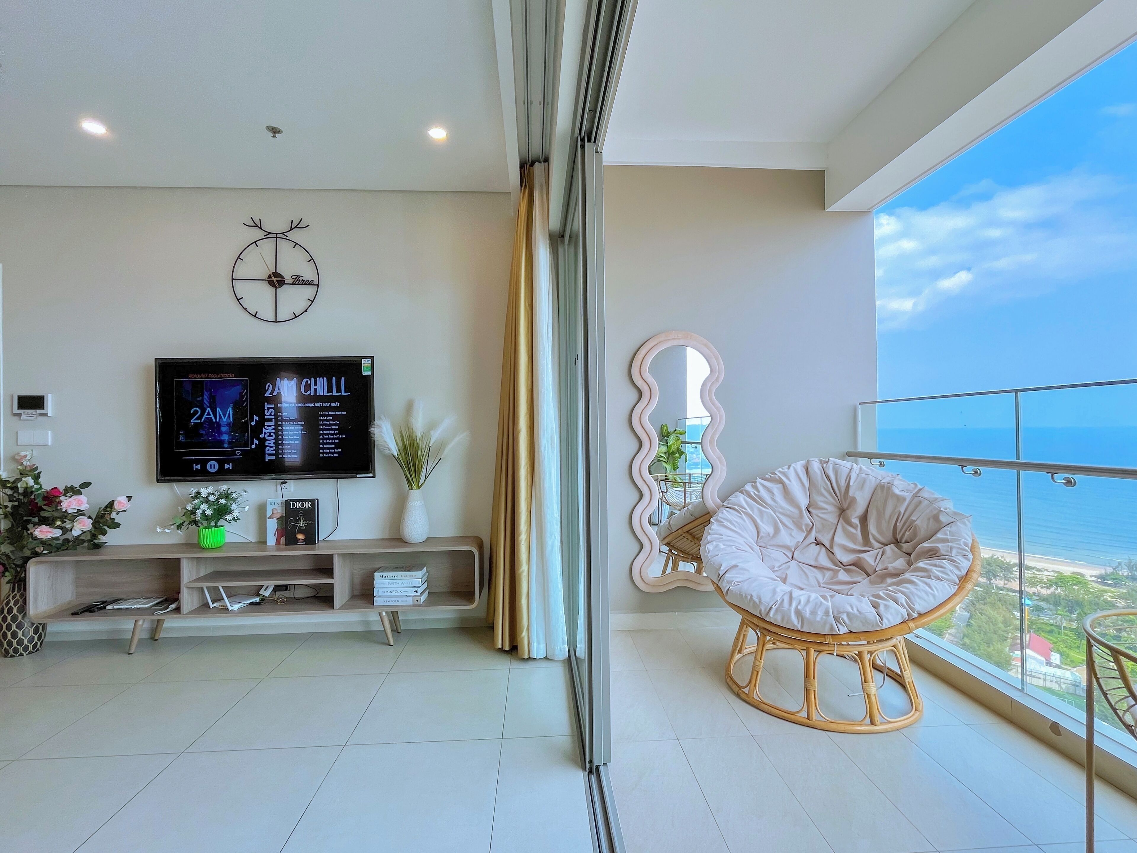 A modern living room with a flat-screen TV, a clock, a console table with books and a vase, and a wicker chair by a large window overlooking the sea.