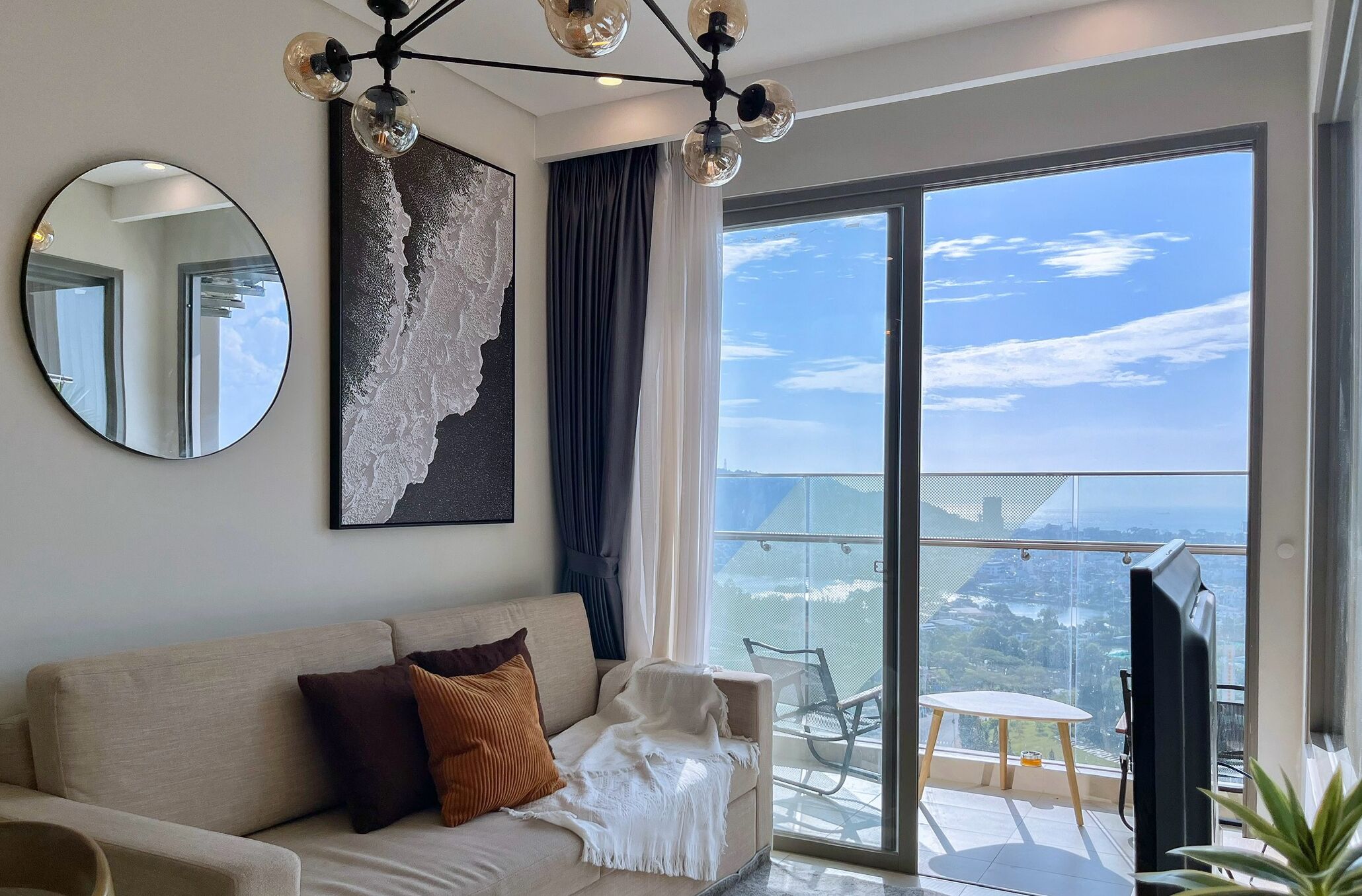 A modern living room with a sofa, a round mirror, a large abstract painting, and a balcony with a view.