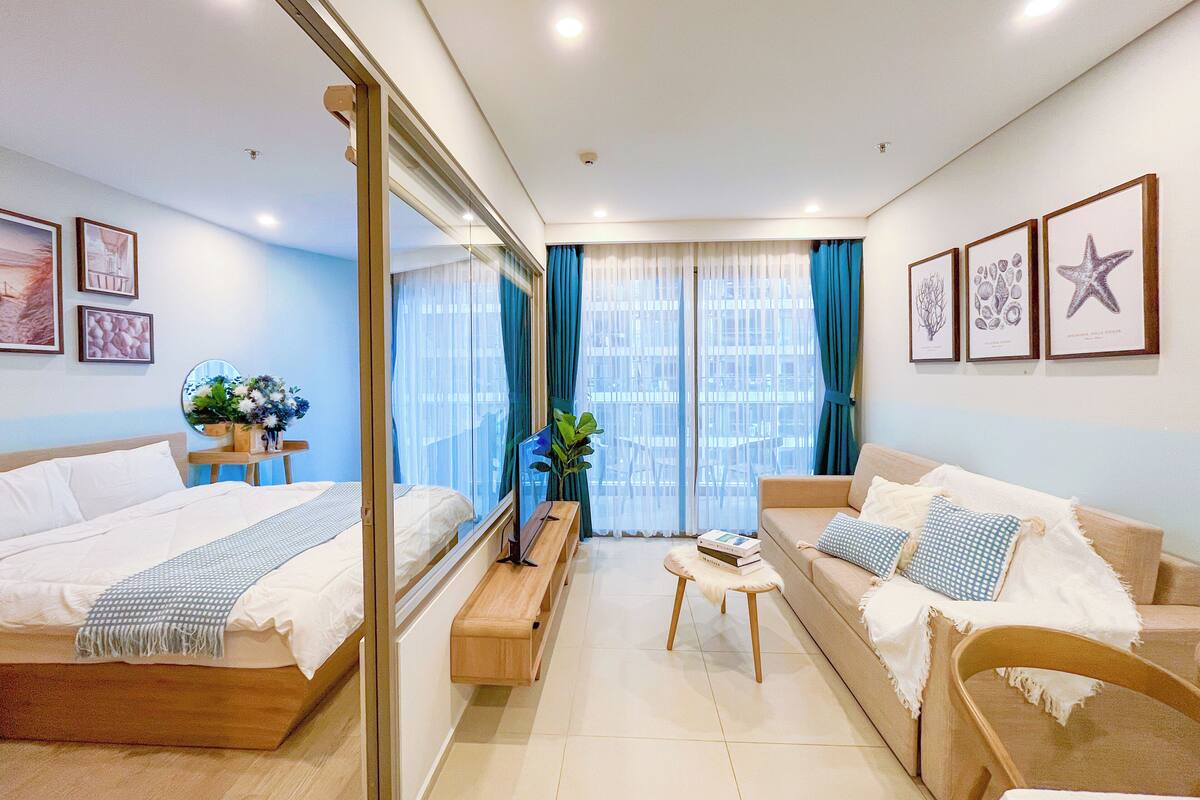 Una habitación de hotel moderna con cama, sofá y una mesita. Hay una ventana con cortinas, un espejo y cuadros en la pared.