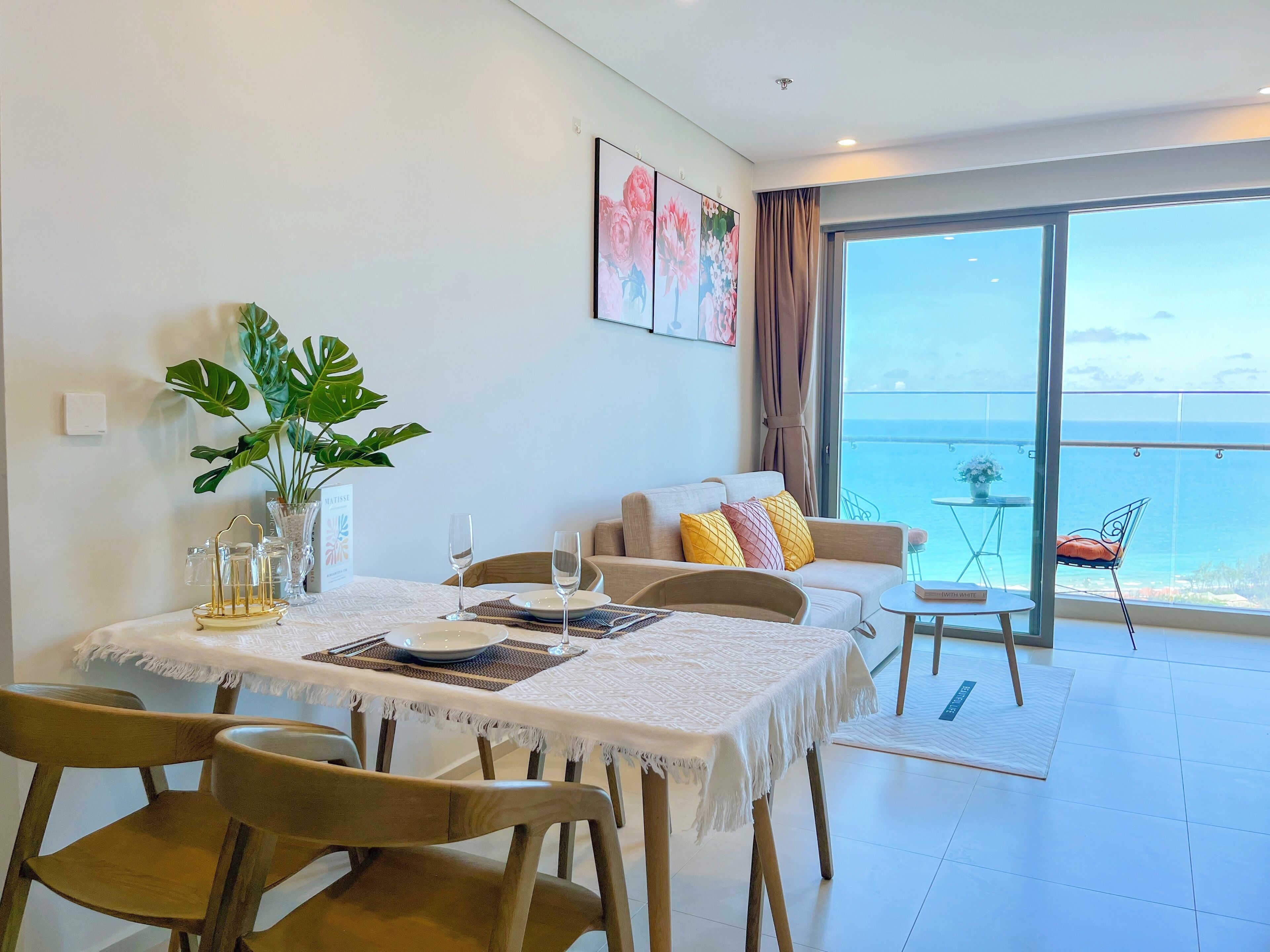 A dining area with a table set for two, a plant, and a view of the sea through large windows.