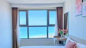 Un cuarto de hotel con un amplio ventanal que ofrece vista al mar, una cama bien tendida con ropa blanca y una mesita con un jarrón de flores rosadas.