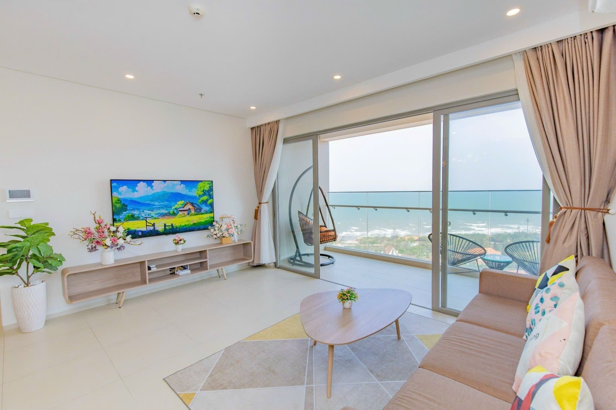 A modern living room with a large TV, a sofa, a coffee table, and a balcony with a view of the sea.