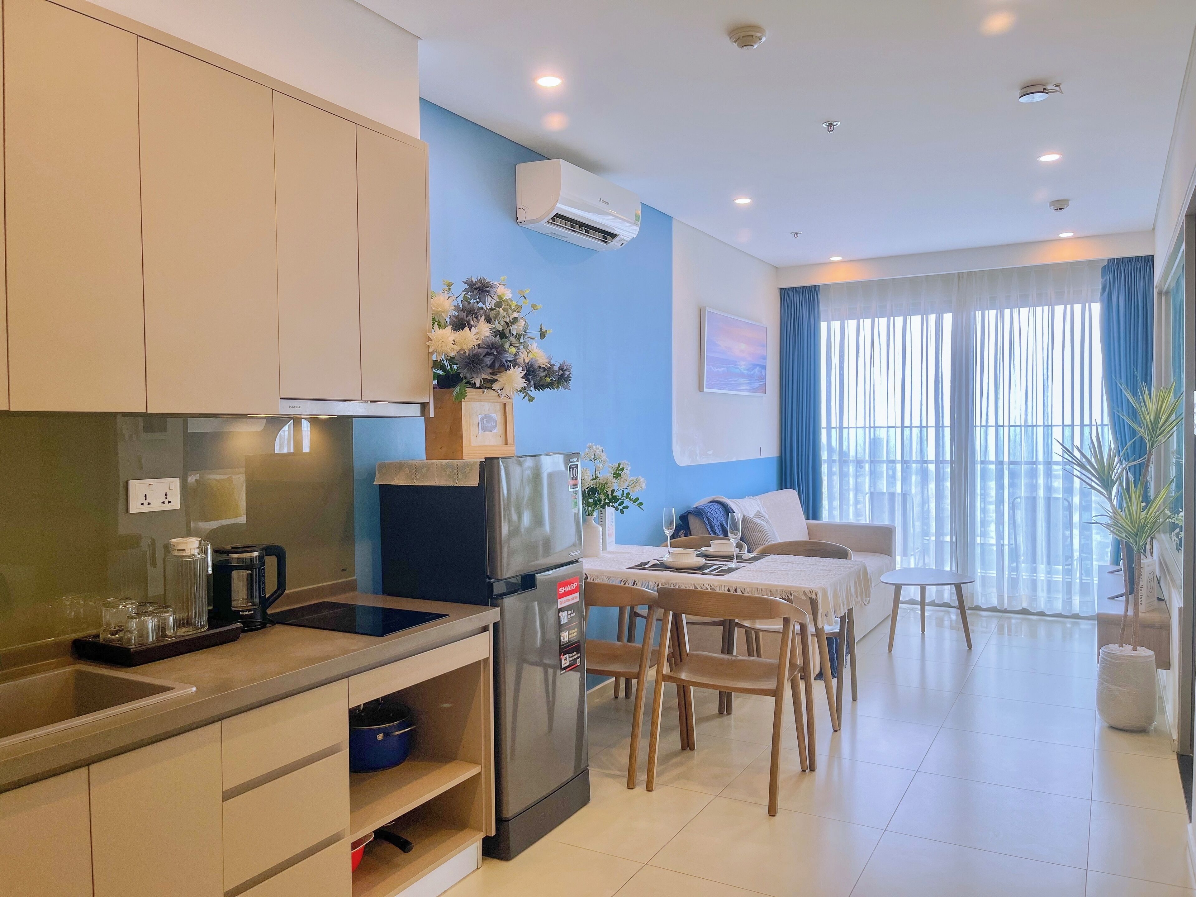A modern kitchen with a refrigerator, stove, and sink. There is a dining area with a table and chairs. The room has a large window with curtains and a view of the outside.