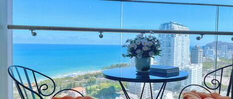 Un balcón con vista al océano y al paisaje urbano, con dos sillas y una mesa con un jarrón de flores.