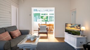 Uma sala de estar com sofá, mesa de centro com bandeja, televisão sobre um suporte e uma área de jantar com mesa e cadeiras.