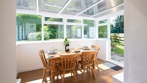 Um solário com uma mesa posta para quatro pessoas, rodeado por grandes janelas com vista para um jardim.