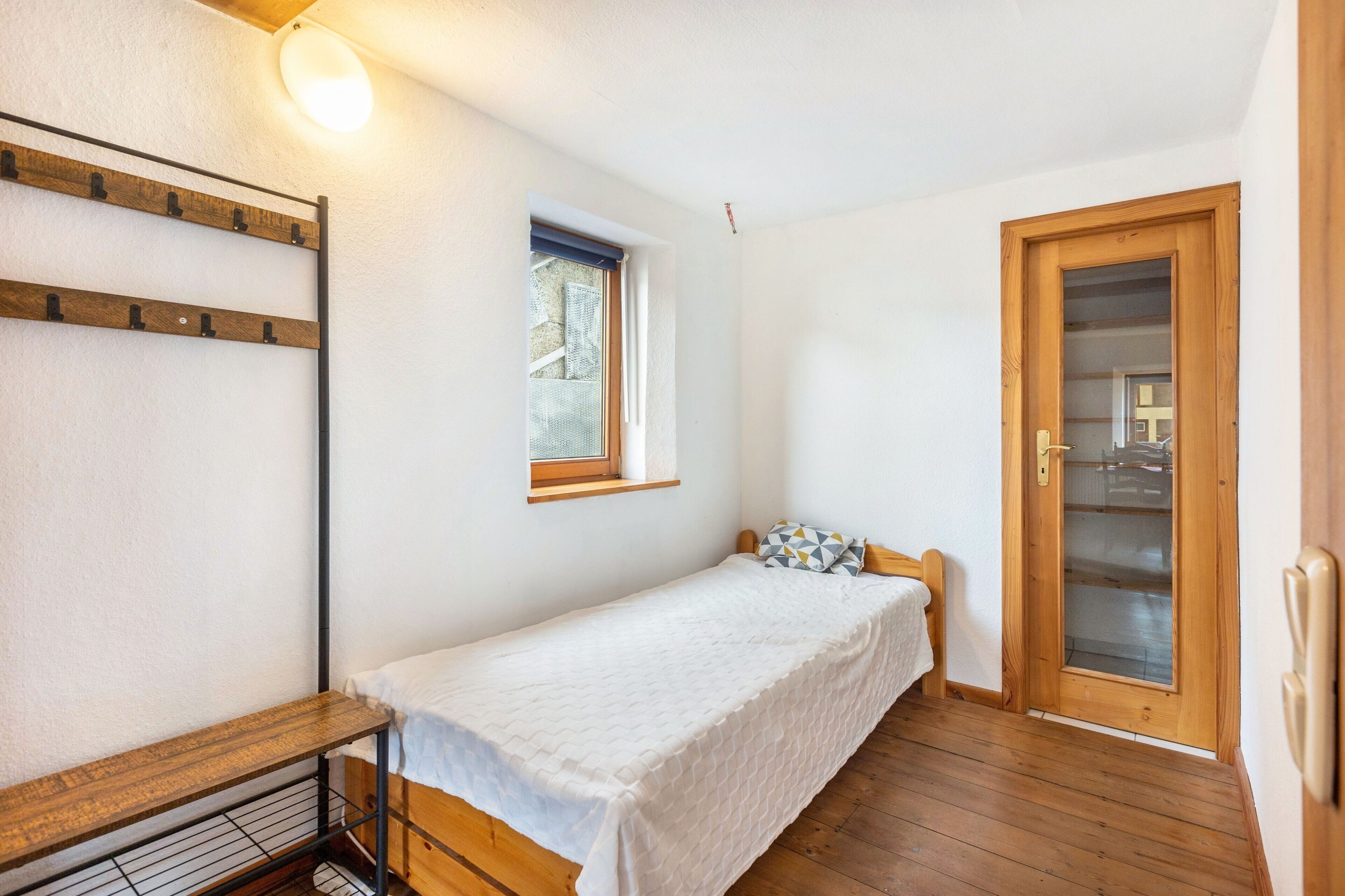 A small, single-bed room with a wooden floor, a window, a door, and a shelf with a lamp.