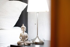 Uma cama com roupa de cama branca, uma pequena estátua de Buda e um abajur em uma mesa de cabeceira.