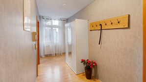 Un pasillo con suelo de madera, un armario blanco, un espejo, un perchero de madera con ganchos y una planta en maceta con flores rojas.