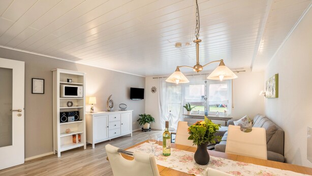A dining area with a wooden table, chairs, a vase with flowers, a television, and a white cabinet with shelves. - Holiday Apartment Odenwald with Terrace and Wi-Fi (Wald-Michelbach)