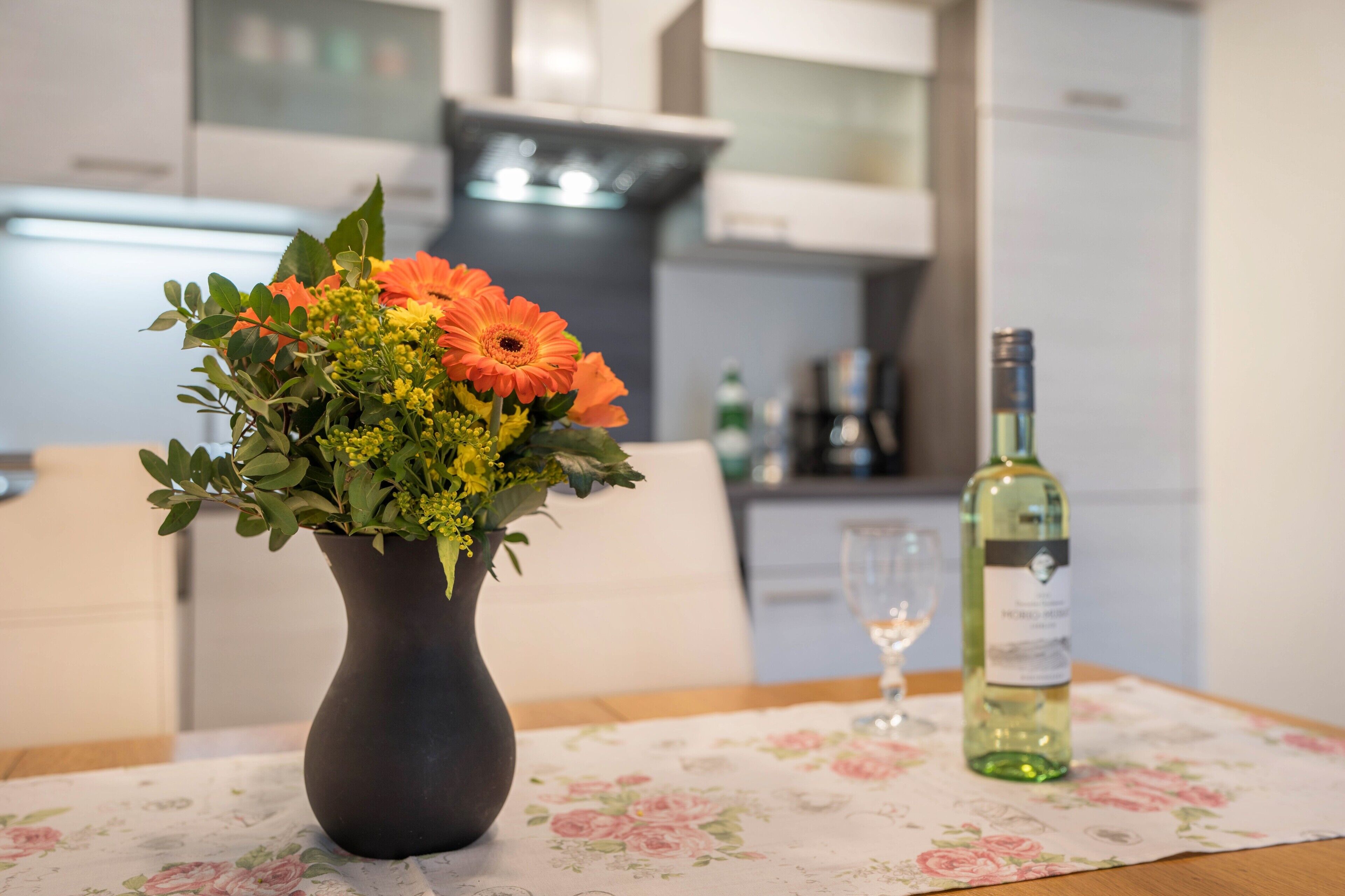 A dining table with a floral tablecloth, a vase with flowers, and a bottle of wine.