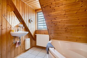 A wooden cabin bathroom with a sink, mirror, window, radiator, and bathtub.