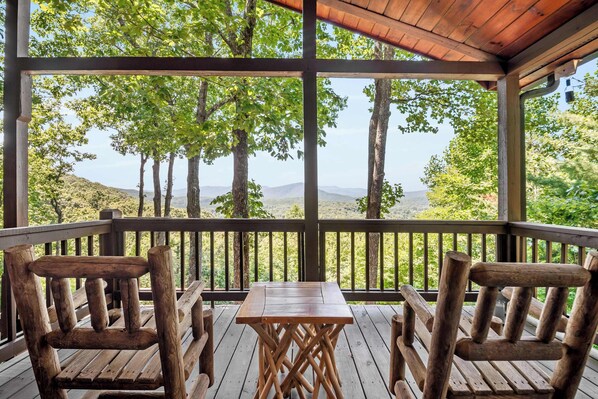 A wooden deck with a table and chairs, overlooking a mountainous landscape with trees.