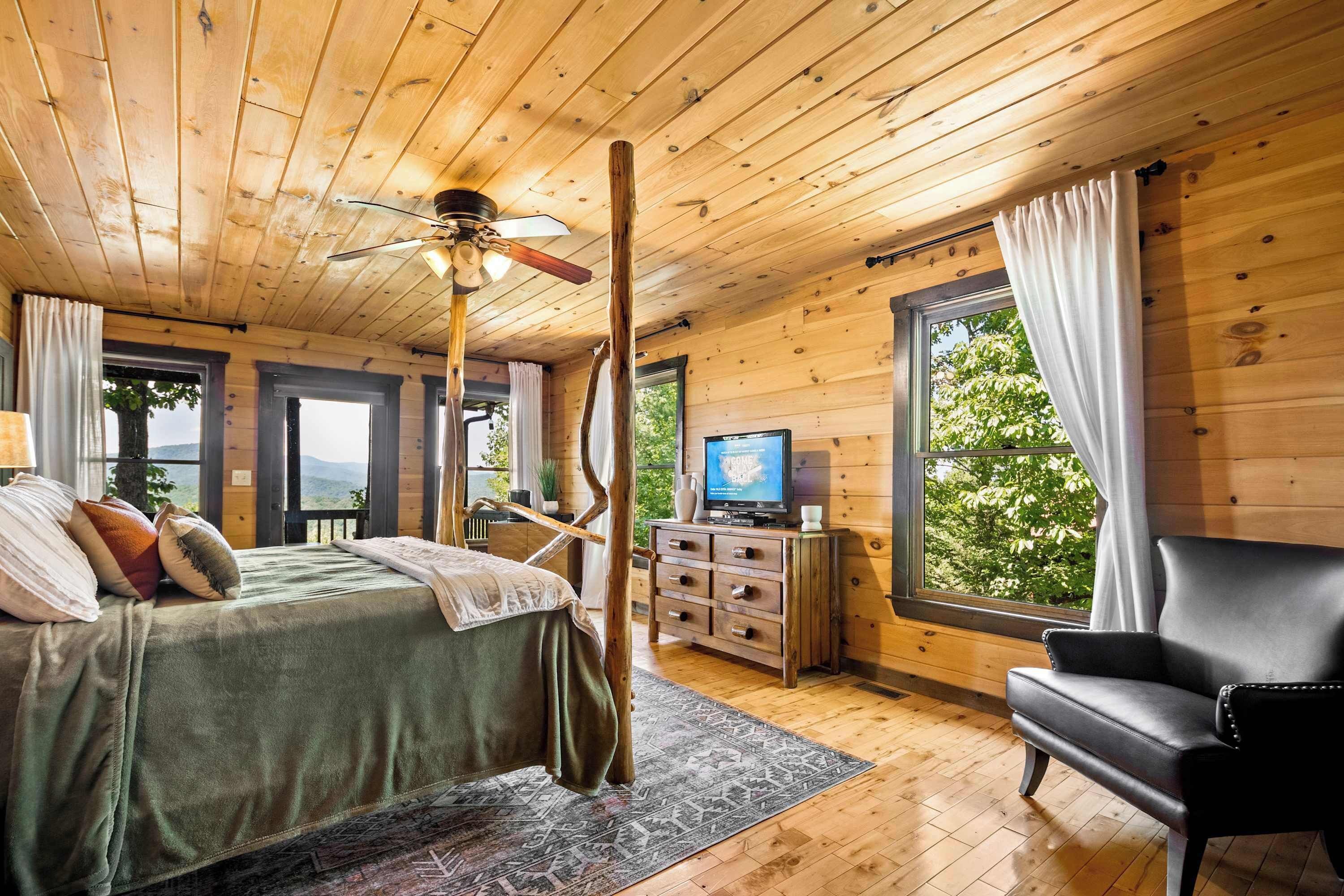 A wooden cabin bedroom with a bed, a chair, a TV, and a window with curtains.