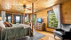 A wooden cabin bedroom with a bed, a chair, a TV, and a window with curtains.