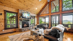 A cozy cabin interior with a stone fireplace, wooden walls, large windows, and a comfortable seating area.
