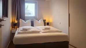 A neatly made bed with pillows and a folded towel, bedside lamps, a window with curtains, and a door with a handle.