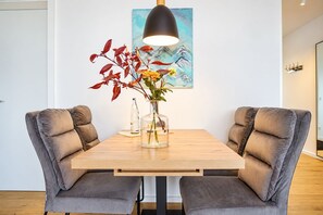 A dining area with a wooden table, two grey upholstered chairs, a vase with flowers, a painting on the wall, and a pendant light.