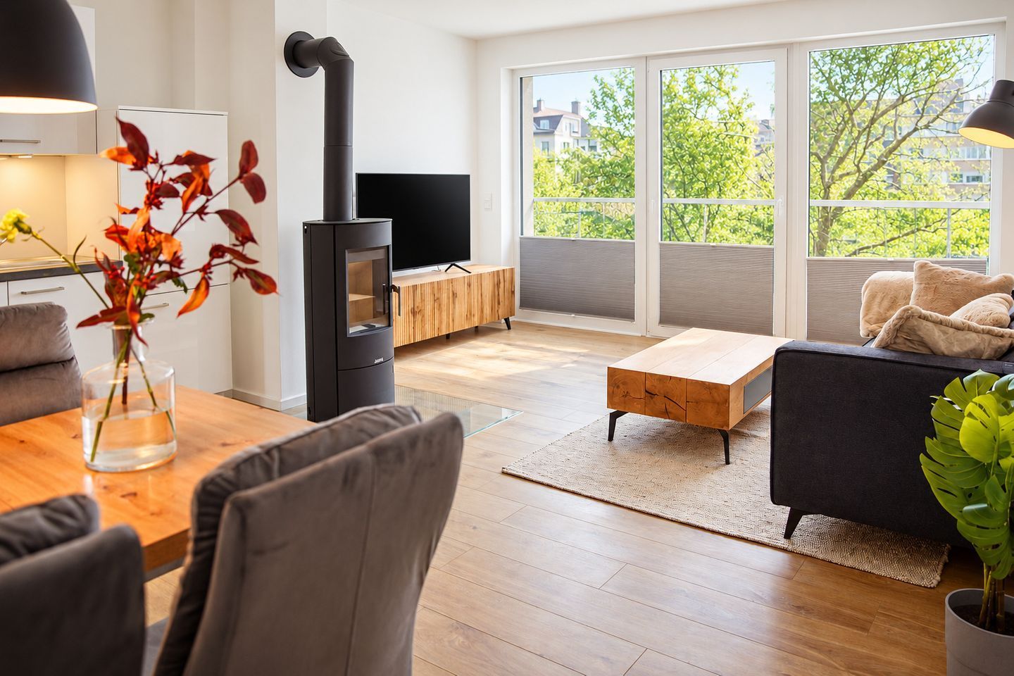 Een moderne woonkamer met een zwart houtkacheltje, een houten eettafel, een bank en een televisie.