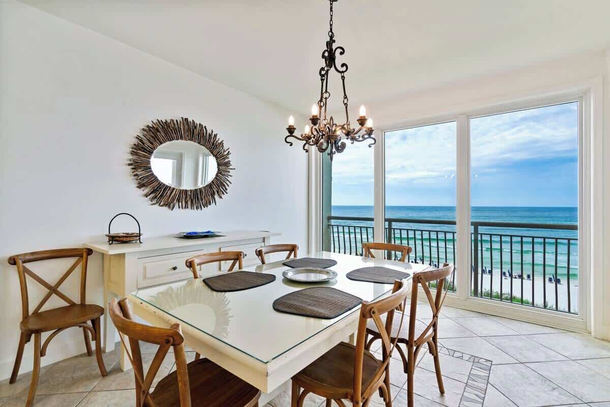 A dining area with a white table, wooden chairs, a chandelier, a mirror, and a view of the ocean.