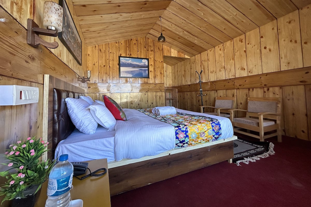 Un dormitorio de cabaña de madera con una cama, una silla, una mesita de noche, una botella de agua y un cuadro en la pared.