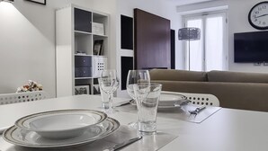 A modern dining area with a white table set for two, a bookshelf, a sofa, and a TV mounted on the wall. - Italianway Comfort - Torino 57 A (Milan)