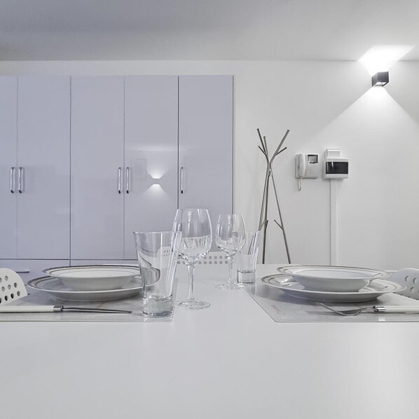 A modern dining area with a white table set for four, including plates, glasses, and cutlery. There are cabinets with handles and a wall-mounted telephone and intercom system. - Italianway Comfort - Torino 57 A (Milan)
