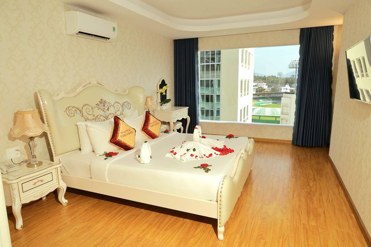 A hotel room with a large bed, a nightstand, a mirror, and a view of a stadium through the window.