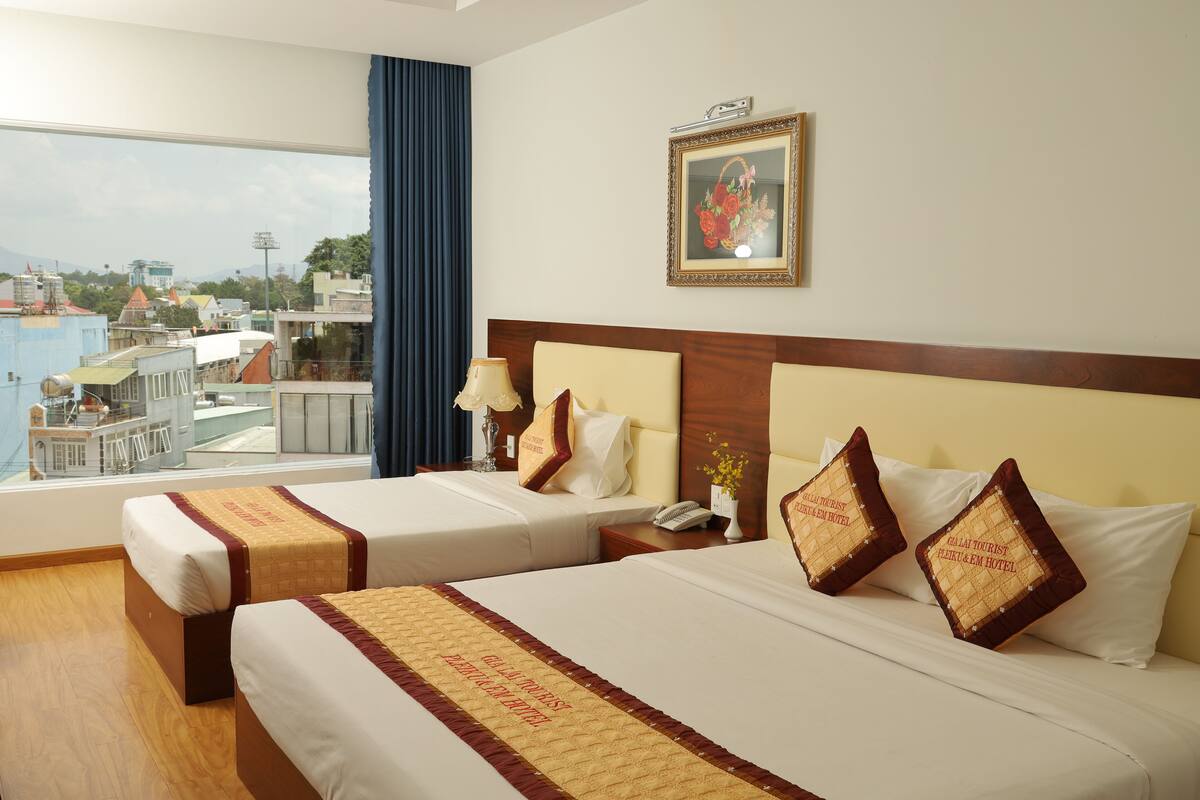 A hotel room with two beds, a large window with a view of buildings, a framed picture on the wall, and a decorative pillow with text.