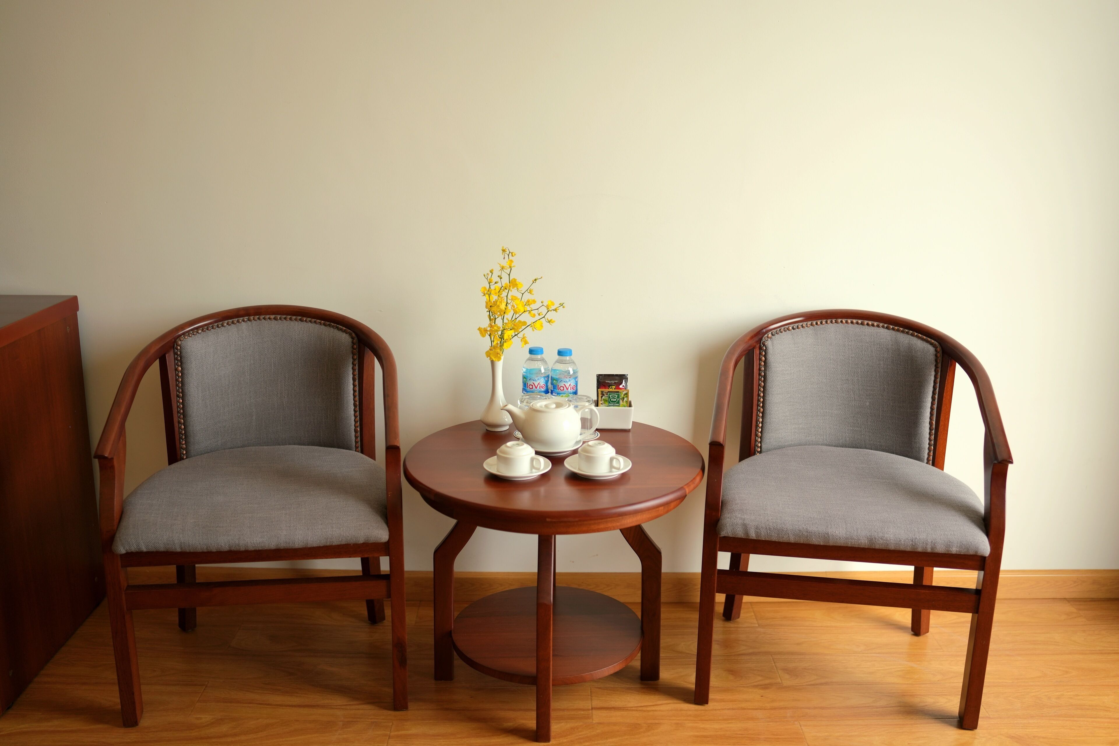A room with two wooden chairs and a small round table with a vase of yellow flowers, a teapot, cups, and bottled water.