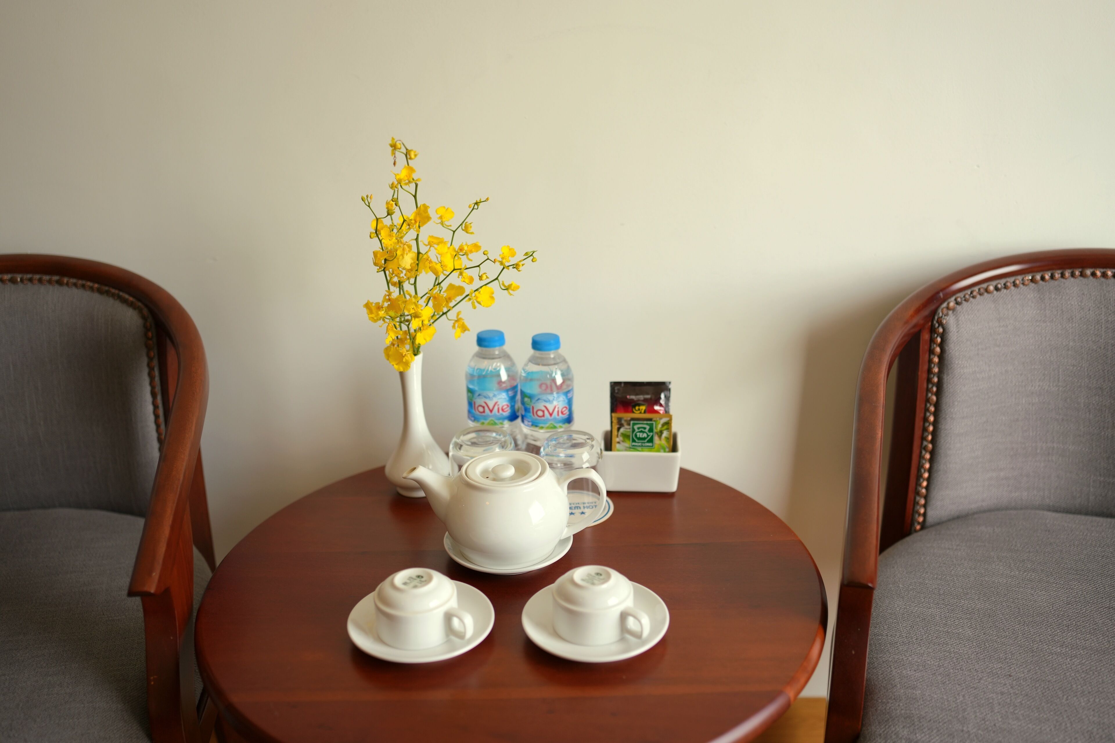 Une table ronde en bois, sur laquelle se trouvent une théière blanche, deux tasses blanches, un vase avec des fleurs jaunes, deux bouteilles d’eau et une petite boîte contenant des sachets de thé.