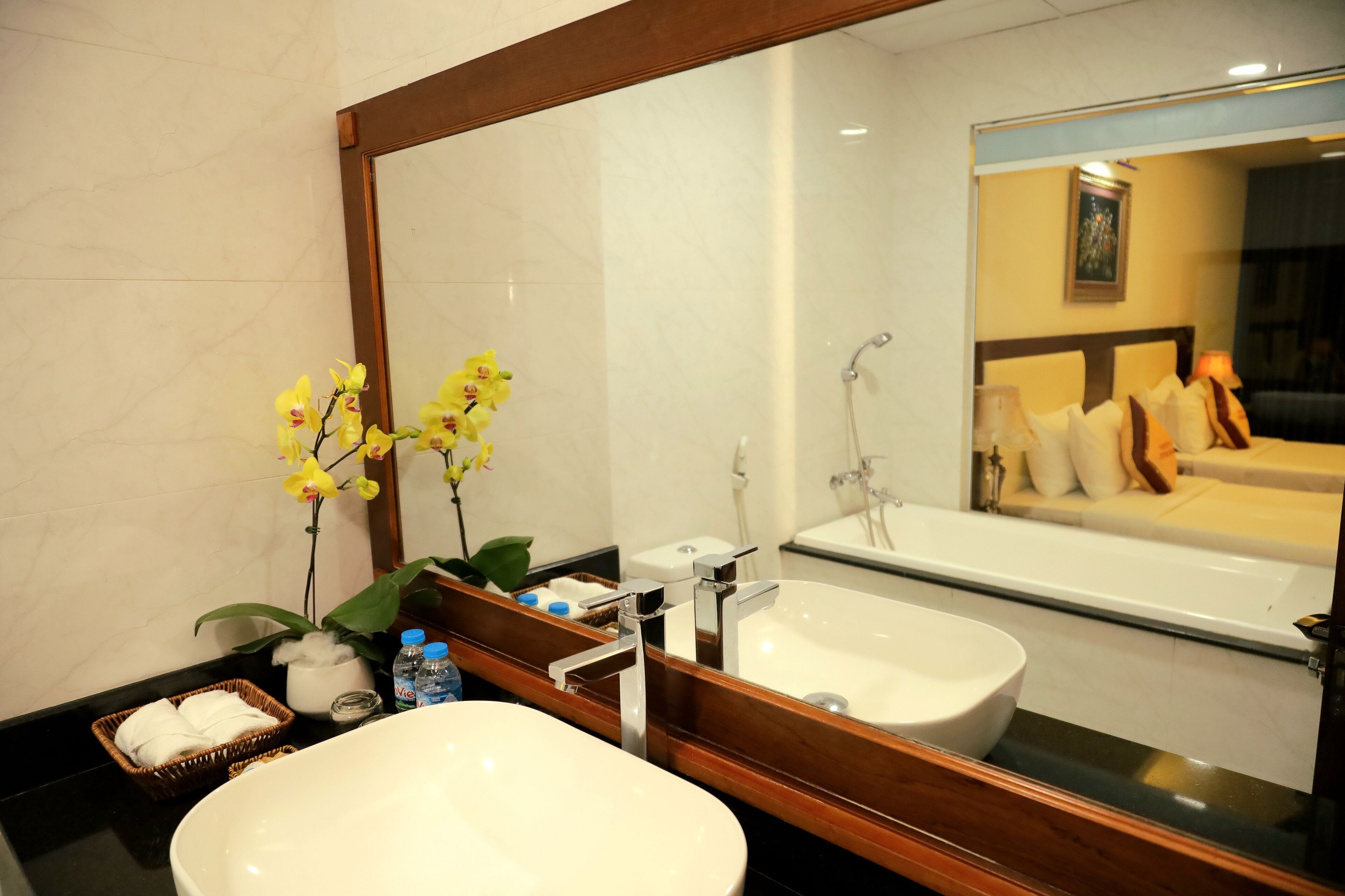 A bathroom with a large mirror, a white sink, and a bathtub. There are yellow orchids on the counter, a basket of towels, and a bottle of water.