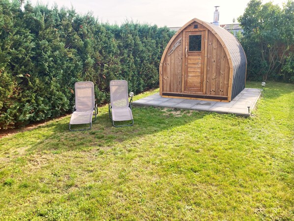 Un abri en bois avec une porte et une petite fenêtre, deux chaises en métal et une zone pavée dans une cour gazonnée.