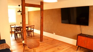 Una habitación con piso de madera, una mesa de comedor con sillas, un sofá, un televisor montado en la pared y un armario de madera.