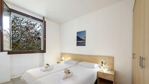Een slaapkamer met een groot raam, een bed met witte lakens, een nachtkastje met een lamp en een houten garderobe.