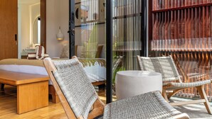 A room with a bed, two wicker chairs, a round table, and a large window with vertical blinds. - Red Sunset Villa by Betterplace (Canggu)
