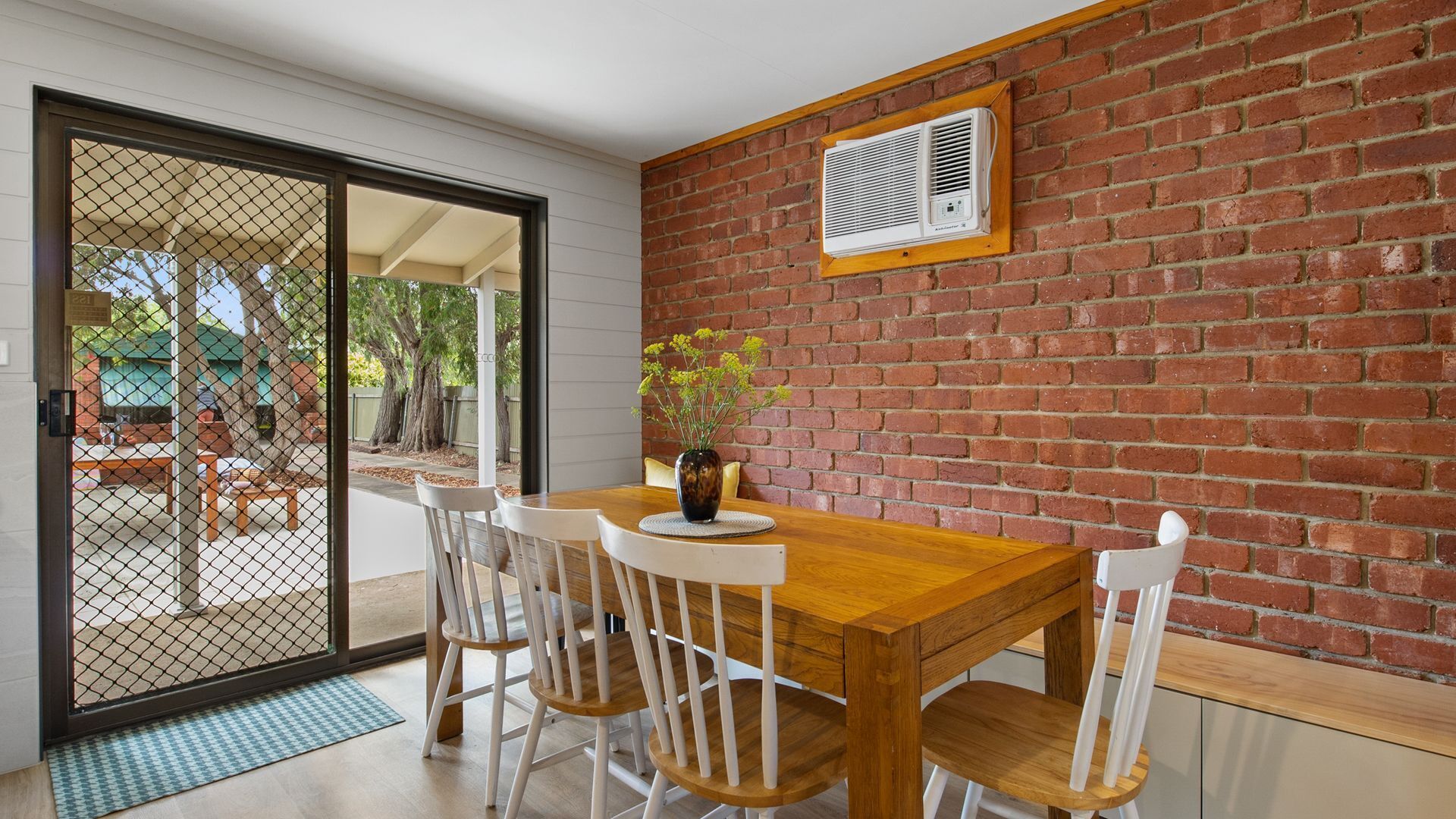 A dining area with a wooden table and white chairs, a brick wall, and a sliding glass door leading to an outdoor patio.