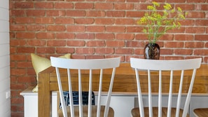Two white chairs with wooden backrests and a vase with yellow flowers on a table against a brick wall.