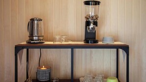 Een houten wand met een metalen plank waarop een koffiezetapparaat, een glazen kan en een kaars staan. Daaronder staan glazen potten en een kandelaar.
