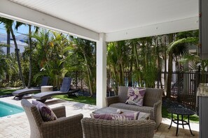 Un espacio exterior techado con mobiliario de mimbre, una piscina y abundante vegetación.