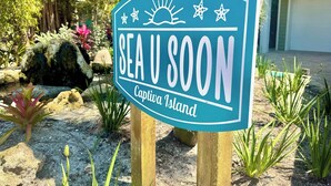 Een bord met de tekst 'SEA U SOON Captiva Island', omringd door tropische planten en een rotsachtig landschap.