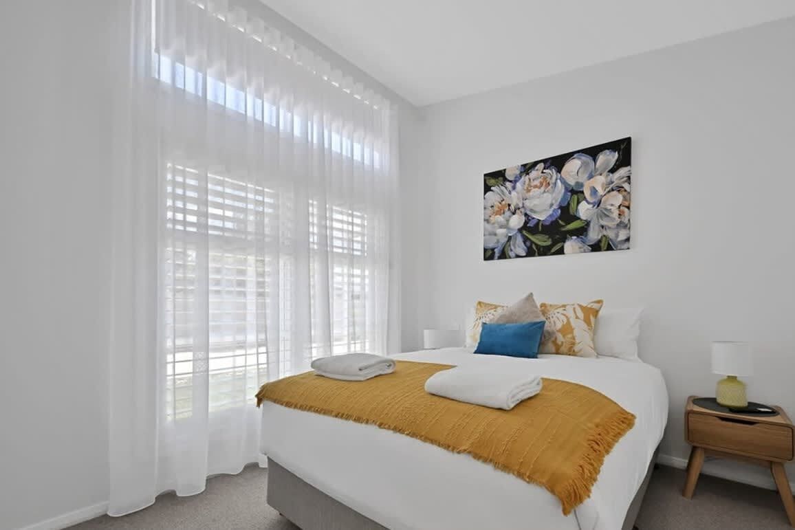 Ein Hotelzimmer mit einem großen Bett, weißen Vorhängen, einem Blumenbild und einem Nachttisch mit einer Lampe.