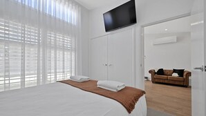 A modern hotel room with a large bed, a flat-screen TV, and a view of a cityscape through sheer curtains. - The Elm Apartments - The Cowhide Room (Orange)