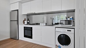 A modern kitchen with white cabinets, a stainless steel refrigerator, a built-in oven, a microwave, and a washing machine. - The Elm Apartments - The Cowhide Room (Orange)