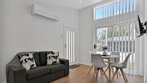 A modern living room with a dark grey sofa, a dining table with chairs, and a large window with vertical blinds.