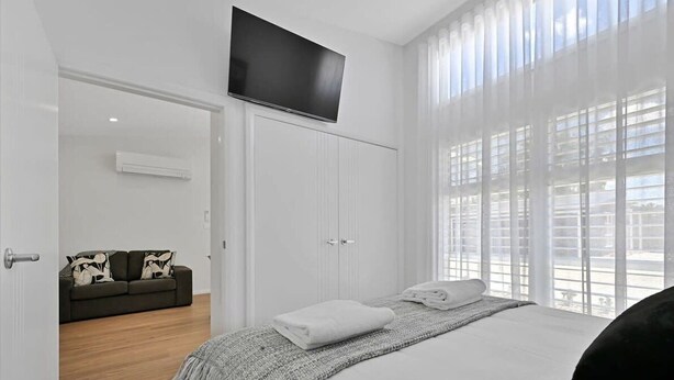 A bedroom with a bed, a TV mounted above it, a sofa in the adjacent room, and a large window with blinds.