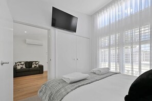 Une chambre à coucher avec un lit, un téléviseur fixé au mur au-dessus du lit, un canapé dans la pièce adjacente et une grande fenêtre avec des stores.